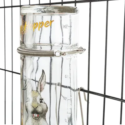 Glass drinking bottle with yellow 'PetHopper' lettering, measuring scale up to 500ml and rabbit motif, attached to a metal grid with holder.