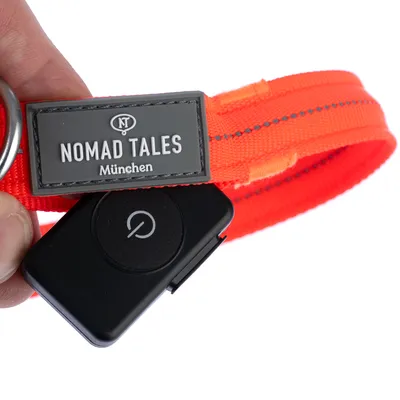 Collier orange avec étiquette grise « NOMAD TALES München » et boîtier noir avec symbole marche/arrêt visible, matériau textile et plastique.