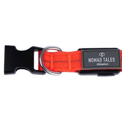 Collier en tissu orange avec boucle plastique noire, anneau métallique et étiquette visible : NOMAD TALES München.
