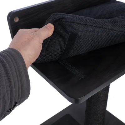 Main soulevant un coussin en tissu noir fixé par scratch sur une plateforme en bois foncé d’un arbre à chat, montrant la fixation amovible du coussin.