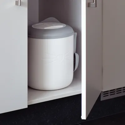 White cat litter container with grey lid, cat face embossing, placed in a cupboard.