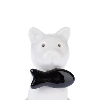 Figurine en céramique blanche en forme de chat tenant un poisson noir dans sa bouche, deux trous visibles sur le visage.