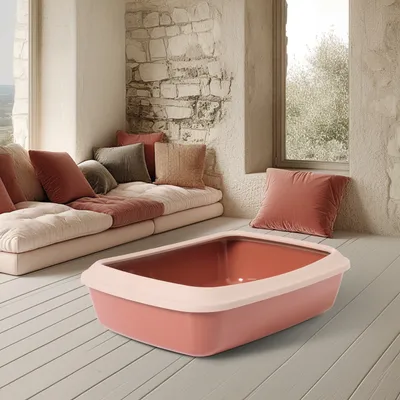 Arenero rosa para gatos con borde color crema, forma rectangular, presentado en una sala de estar con cojines beige y rojos en un banco.