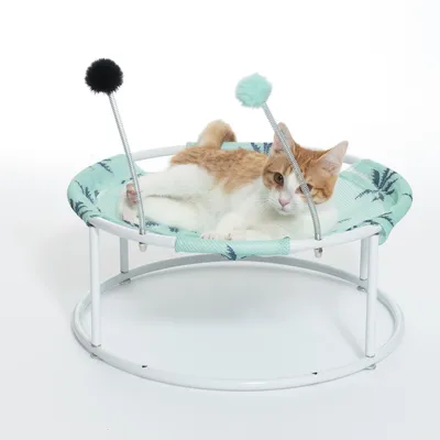 Katzenliege mit rundem Metallgestell, grünem Stoffbezug und zwei Spielstäben mit schwarzen und grünen Plüschbällen. Katze liegt entspannt auf der Liege.