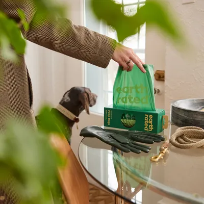 Earth Rated dog waste bags in green packaging, person taking out a bag. Accessories such as gloves and lead visible on table.