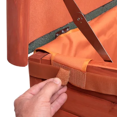 Nahaufnahme eines orangefarbenen Klettverschlusses an einem Holzrahmen, der von einer Hand geöffnet wird. Metallstreben und Stoffpolsterung sichtbar.