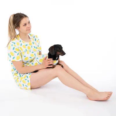Femme assise portant un pyjama à motifs citrons assorti à un petit chien noir et marron vêtu du même motif, sur fond blanc.