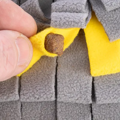Una mano mostra come una leccornia viene inserita nella tasca di stoffa gialla di un tappeto olfattivo grigio.