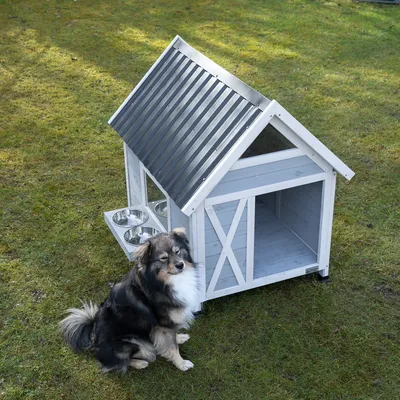 Gråt hundehus med metaltag og to integrerede foderskåle. Hund sidder ved siden af på en græsplæne.