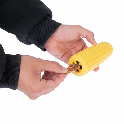 Giocattolo giallo a forma di pannocchia di mais, riempito con uno snack. Mani mostrano come inserire lo snack.