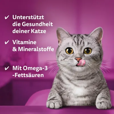 Besedilo v nemščini: Unterstützt die Gesundheit deiner Katze, Vitamine & Mineralstoffe, Mit Omega-3-Fettsäuren. Siva mačka z roza jezikom na vijoličnem ozadju.