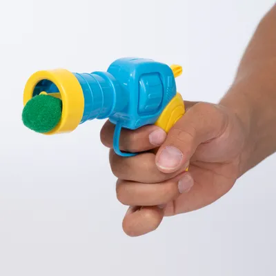 Hand hält eine blaue und gelbe Spielzeugpistole mit grünem Ball vorne, geeignet für interaktives Spielen.