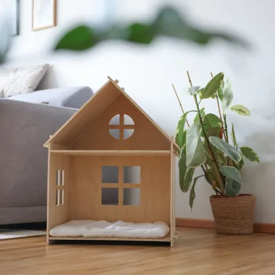 Wooden house with window design and soft insert, suitable as pet accommodation. Placed in a living room next to a plant.