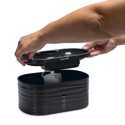Fontaine à eau noire en plastique beeztees, couvercle amovible tenu par une main, réservoir et filtre visibles à l’intérieur.