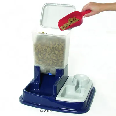 Distributeur automatique de croquettes en plastique bleu et gris, couvercle ouvert, main versant des croquettes avec une pelle rouge, deux compartiments pour nourriture et eau visibles.