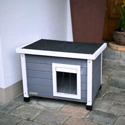 Grey wooden dog house with white frame, black roof and transparent door flap. Standing on stone floor in front of a house wall.