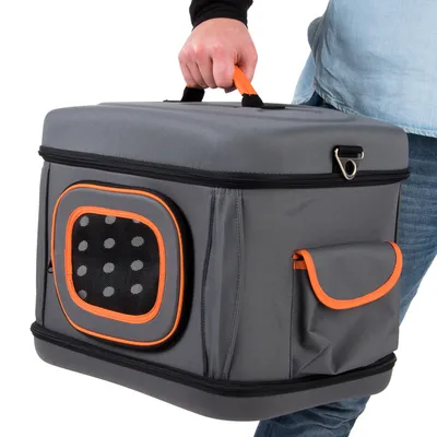 Sac de transport rigide gris avec poignée orange, poche latérale et fenêtre d’aération à pois, porté à la main par une personne en jean et chemise claire.