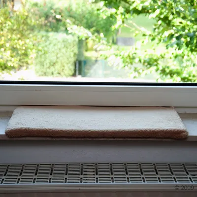 Coussin rectangulaire beige en tissu posé sur un rebord de fenêtre au-dessus d’un radiateur, sans marque visible ni texte.