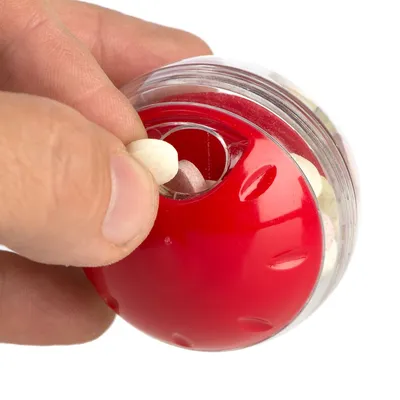 Mano che inserisce una pastiglia in una palla rossa trasparente riempita di snack o croccantini. Materiale visibile: plastica.