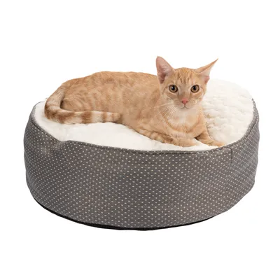 Rundt, polstret kæledyrsseng med gråt prikket mønster og hvid plyssoverflade. En orange korthåret kat ligger på den.