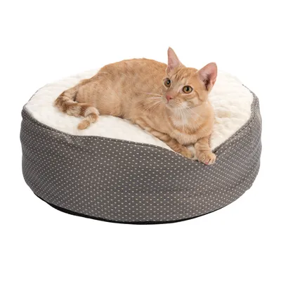 Rundt, polstret kæledyrsseng med gråt stof og hvide prikker, overside af hvid plys. Kat ligger afslappet på sengen.