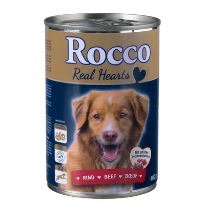 Rocco Real Hearts tinned dog food, 400g. Visible text: Rind, Beef, Bœuf. Packaging shows icons for extra large pieces, fresh ingredients, and 100% meat content.