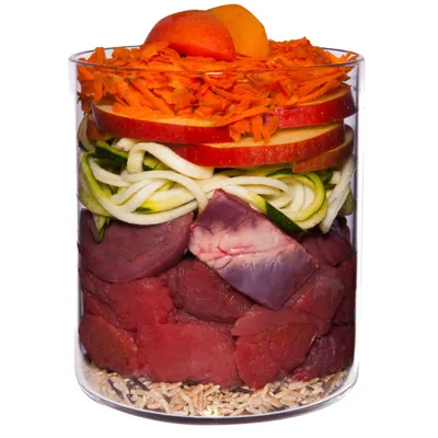 Glass container with visible layers of raw beef chunks, organ meat, courgette spirals, apple slices, grated carrot, apricot halves and brown rice at the bottom. No brand or text shown.