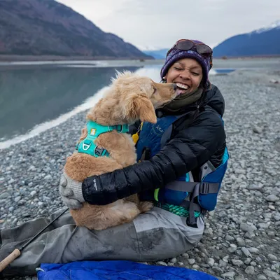 Cane con pettorina turchese seduto in braccio a una persona su una riva di lago ghiaiosa, indossando abbigliamento outdoor e guanti. Pettorina visibile con fibbie e anelli metallici.