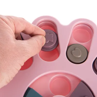 Hand bewegt lila Spielknopf mit Pfotenmotiv auf rosa interaktivem Spielzeug. Fördert Geschicklichkeit und Beschäftigung.
