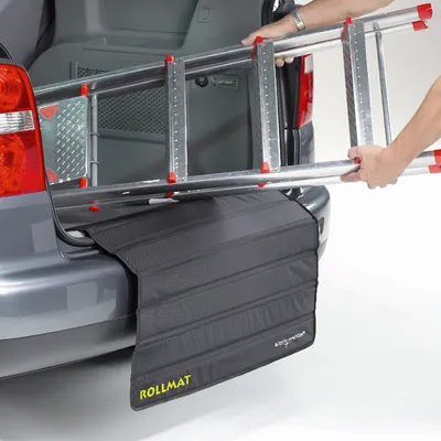 ROLLMAT beskyttelsesmåtte monteret på bilens bagklap, person lægger metalstige ind i bagagerum. Synlig tekst: ROLLMAT, Auto Protect.