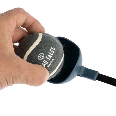 Roka položi siv teniški žogico z napisom 'Mad Tales München' v modro napravo za metanje žogic. Roka položi siv teniški žogico z napisom 'Mad Tales München' v modro napravo za metanje žogic.