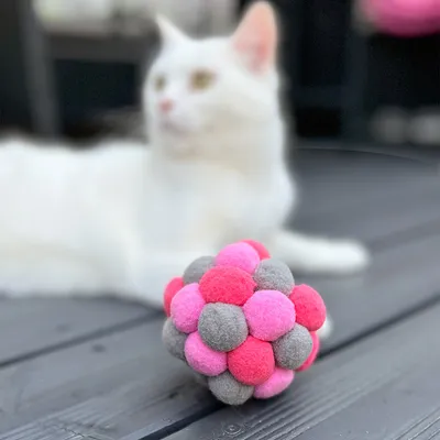 Топка за игра за котки от цветни филцови топчета в розово, сиво и червено. На заден план неясна бяла котка.