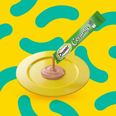 Creamy treat being squeezed from a green Dreamies Creamy sachet onto a yellow plate, showing product texture and serving suggestion.