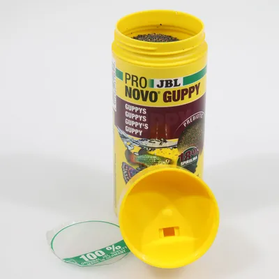 JBL Pro Novo Guppy Fischfutter, gelbe Dose mit geöffnetem Deckel, enthält 18% Spirulina und präbiotische Inhaltsstoffe. Geeignet für Guppys. Siegel: 100% garantiert.