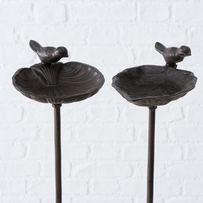 Dwie poidełka dla ptaków z ciemnego metalu z dekoracyjnymi figurkami ptaków na stojakach, odpowiednie do ogrodu lub na zewnątrz.