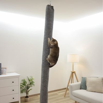Krabpaal in zuilvorm met grijze stofbekleding, kat klimt omhoog. Woonkamer met bank, lamp en commode op de achtergrond.