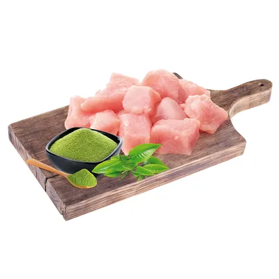 Färska bitar av kycklingkött på träbräda, bredvid skål med grönt pulver och teblad. Ingredienser för djurfoder.