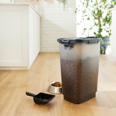 Transparenter Futterbehälter mit schwarzem Deckel, gefüllt mit Trockenfutter. Schwarze Futterschaufel und silberner Napf mit Trockenfutter daneben.