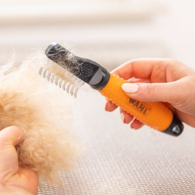Hand houdt WAHL vachtkam met oranje handgreep, verwijdert losse haren uit dichte vachtplukken.