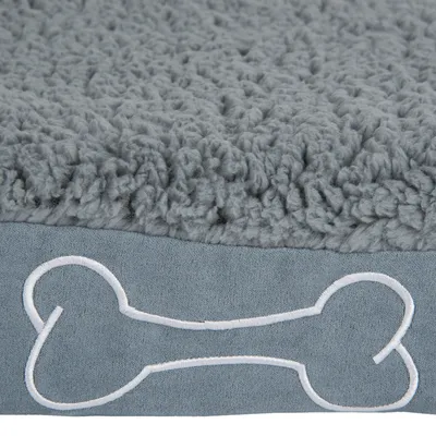 Detalle de cama gris para perro con superficie superior de tela mullida y borde inferior decorado con un dibujo de hueso blanco bordado.