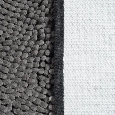 Gros plan sur une surface de tapis en tissu gris à texture chenille et un dessous antidérapant blanc à motifs triangulaires, séparés par une bordure noire.
