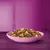 Bowl filled with mixed dry pet food pieces in various shapes and colours, including brown, yellow, green, and orange, on a purple surface with a purple background.