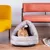 Graues, dreieckiges Katzenbett mit weichem Stoff und hängendem Spielball. Bengal-Katze sitzt darin. Möbel im Hintergrund: gelber Sessel, orangefarbene Bank.