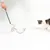 Chat observant une canne à jouer noire avec un long jouet en fausse fourrure grise et plumes, tenue par une main humaine. Jouet pour chat visible sur fond blanc.