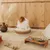 Dog sitting in a round wicker bed on carpet. Next to it a wooden bowl holder with two stainless steel bowls and a rope toy.