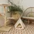 Kratzbaum aus Holz mit drei gepolsterten Ebenen und Spielzeug, daneben ein cremefarbenes Tipi-Zelt mit Katze darin. Dekorative Umgebung mit Pflanzen und Rattanmöbeln.