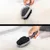 Brosse double face pour animaux, côté picots et côté poils noirs, utilisée sur un pelage blanc. Manche gris visible tenu par une main.