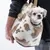 Petit chien blanc dans un sac de transport en tissu à motif tacheté beige et blanc, porté à l’épaule par une personne. Étiquette visible : K9.