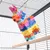 Jouet suspendu en forme de pinata colorée dans une cage à barreaux métalliques, perchoir en bois visible au premier plan.
