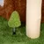 Juguete verde en forma de árbol con base de plumas, sobre alfombra verde junto a un tronco de madera.